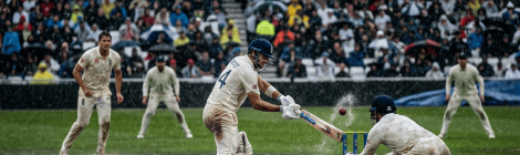 Cricket batter striking the ball as wicketkeeper lunges in heavy rain